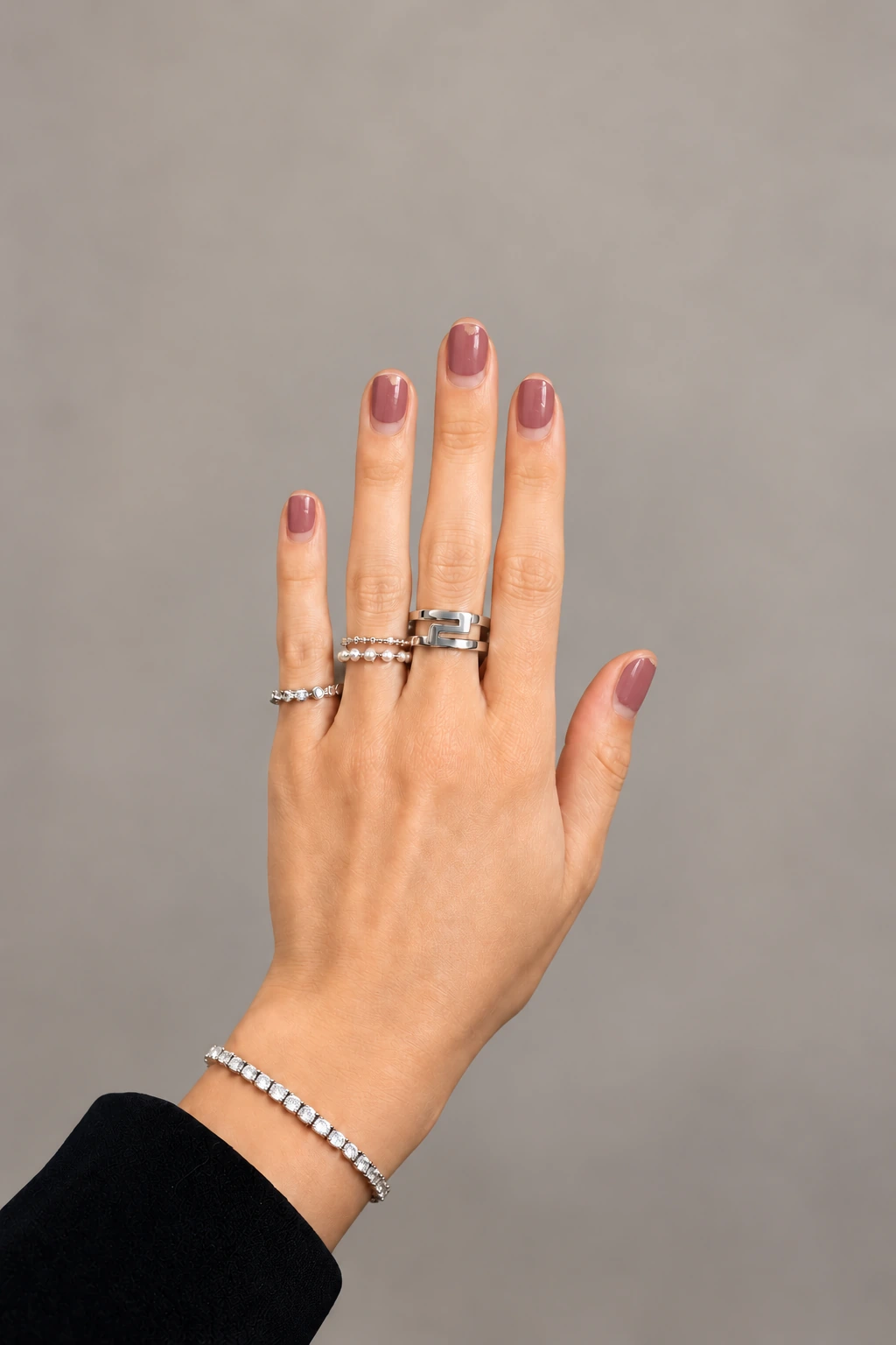A hand wearing silver rings and a bracelet, with short nails and lightly worn mauve polish against a soft neutral background